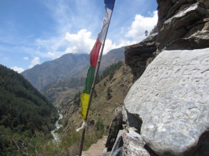 Carved stone on mani wall, prayer flag, and trail far below, reentering the Lower Tsum Valley
